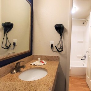 A bathroom with a sink, mirror, hair dryer, and a shower area. - Travelodge Newport News (Newport News)