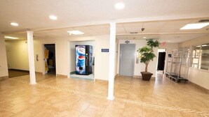 A spacious lobby with a tiled floor, white pillars, a vending machine, a potted plant, and a metal cart.