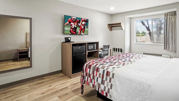 A hotel room with a bed, a desk, a TV, a mirror, and a window with curtains. - Regency Inn & Suites DFW (Euless)