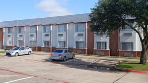 A multi-story building with a brick facade and white trim. There are two cars parked in front, and a tree is visible to the right.