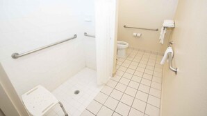 A small, clean bathroom with white tiles, a shower with a handheld showerhead, a toilet, and a grab bar near the toilet. - Regency Inn & Suites DFW (Euless)