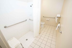 A small, clean bathroom with white tiles, a shower with a handheld showerhead, a toilet, and a grab bar near the toilet. - Regency Inn & Suites DFW (Euless)