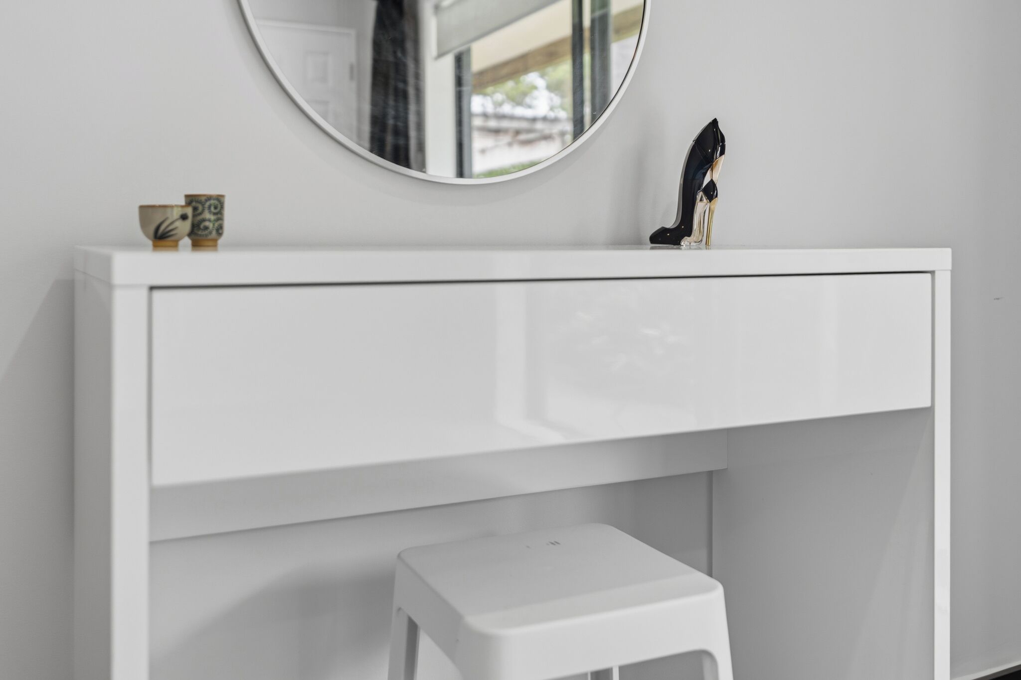 A white vanity with a round mirror, a small stool, and a pair of black high-heeled shoes on top.