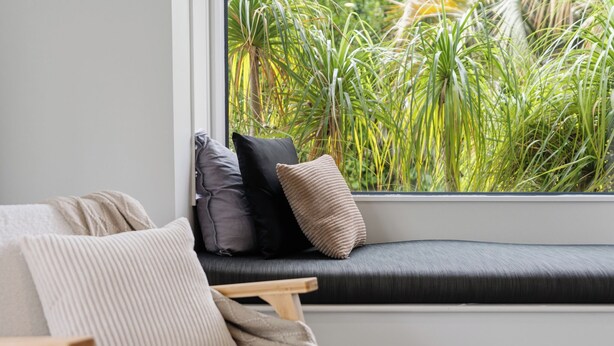 A window seat with cushions, a view of green plants outside.