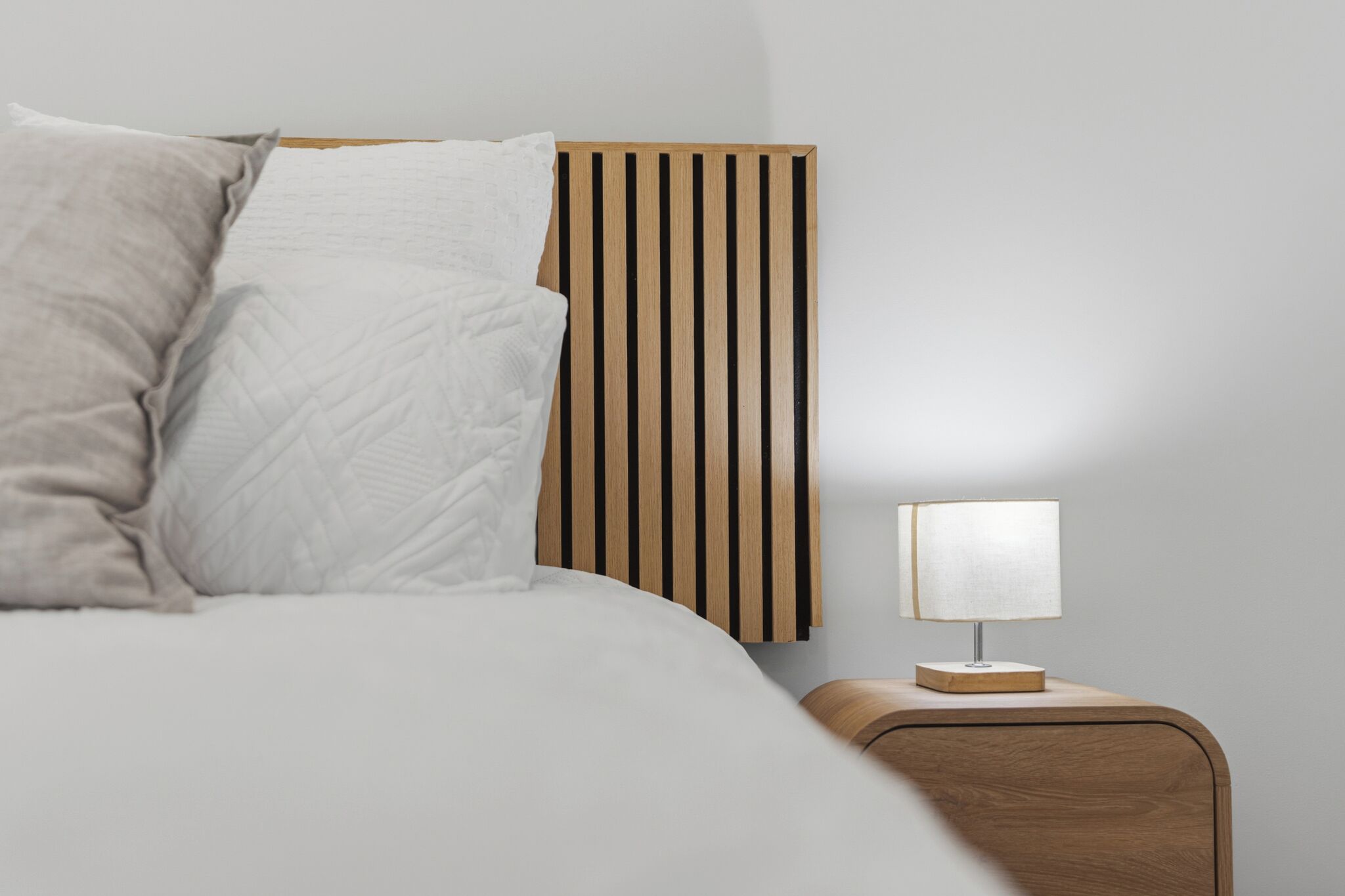 Cama bem arrumada com cabeceira de madeira, mesa de cabeceira com abajur e parede lisa.