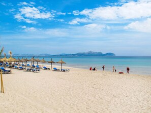 Ein Strand mit Liegestühlen und Reetdach-Sonnenschirmen, einem klaren Himmel und ruhiger See.
