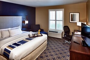 A hotel room with a large bed, a desk with a television, an air conditioning unit, and a window with blinds. - Nationwide Hotel And Conference Center (Lewis Center)