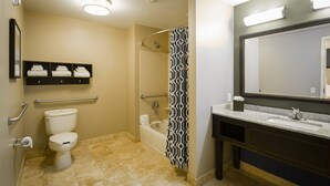 A bathroom with a toilet, bidet, bathtub, and a sink with a mirror. There are towels on a shelf and a shower curtain with a geometric pattern. - Nationwide Hotel And Conference Center (Lewis Center)