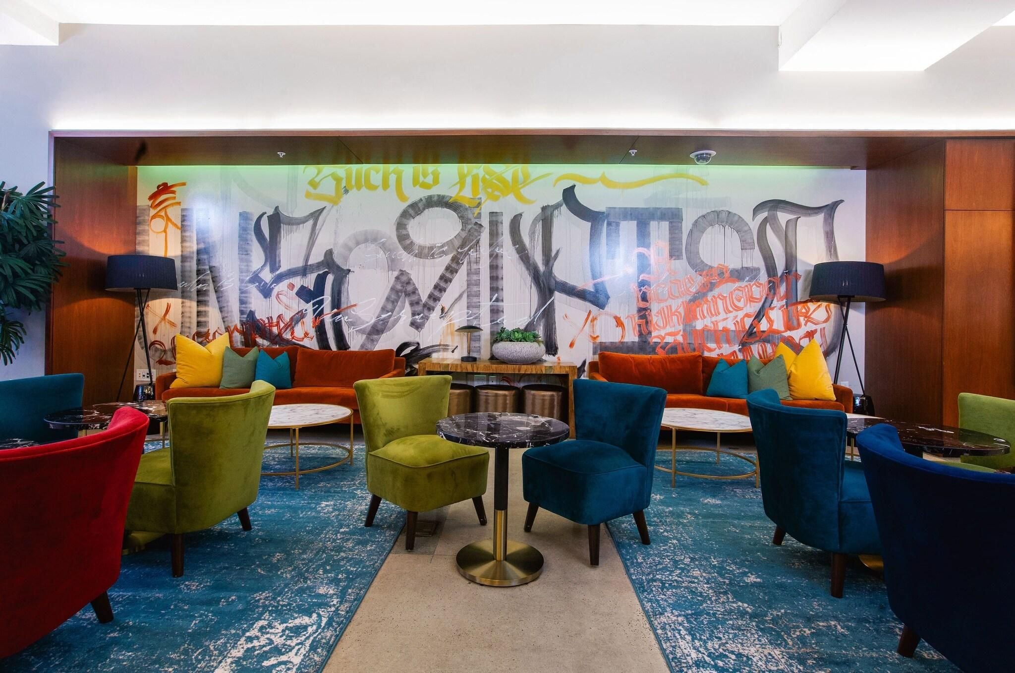 A lobby with colorful chairs, a round table, and a large abstract mural on the wall.