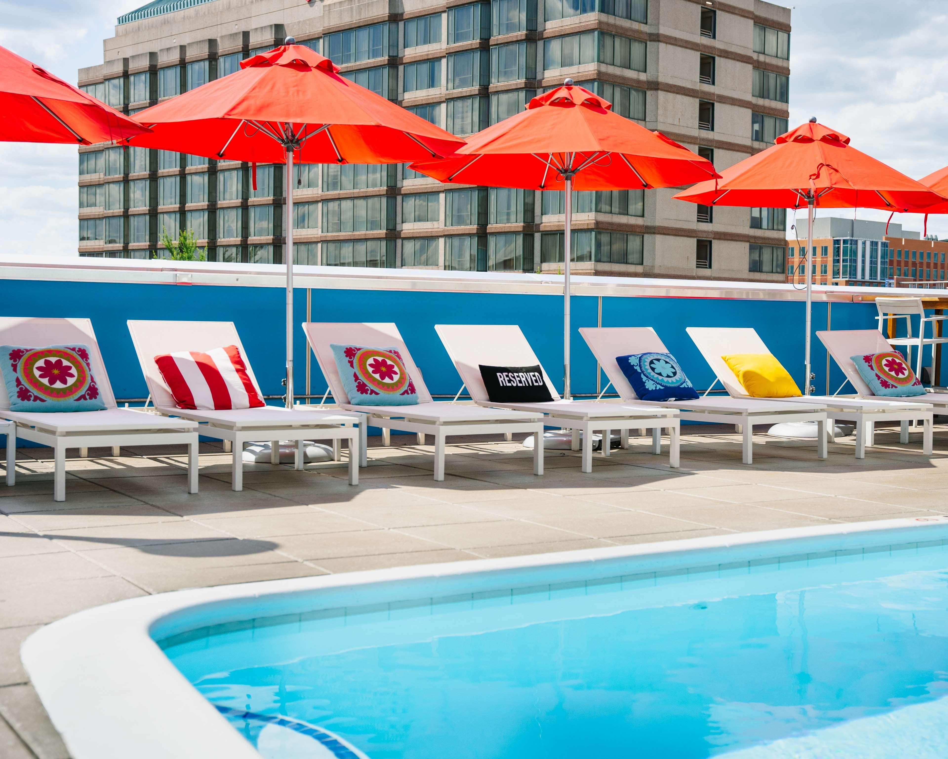 Zona junto a la piscina con sillones de descanso y sombrillas rojas, una pared azul y un edificio al fondo.