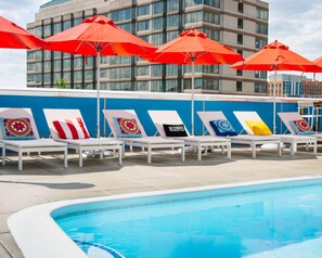 Zona junto a la piscina con sillones de descanso y sombrillas rojas, una pared azul y un edificio al fondo.