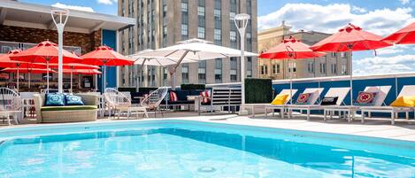 A swimming pool with lounge chairs and umbrellas, a tall building in the background, and a clear sky.