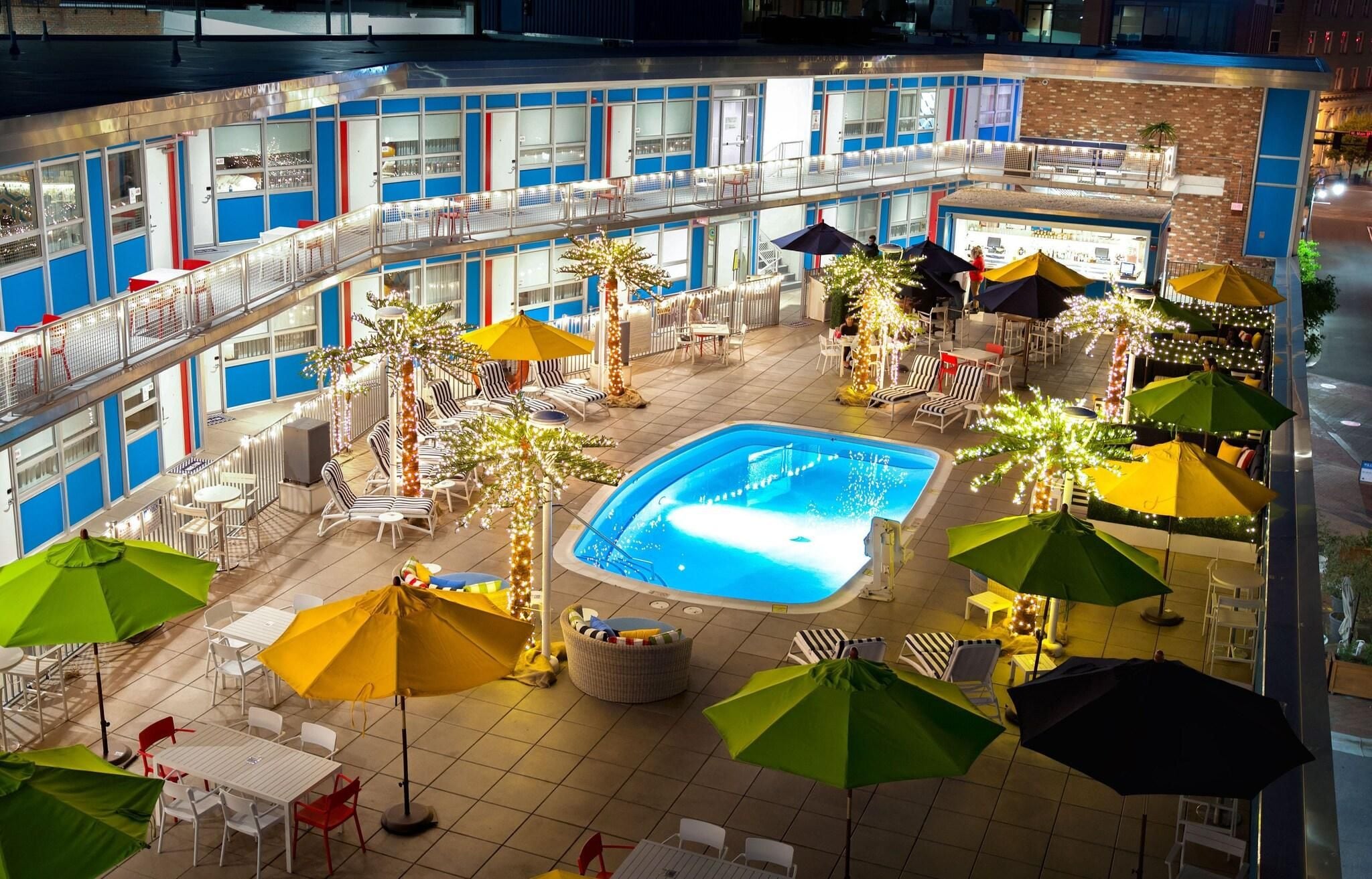 An indoor pool area with multiple lounge chairs and umbrellas, surrounded by a multi-level building with balconies.