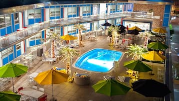 An indoor pool area with multiple lounge chairs and umbrellas, surrounded by a multi-level building with balconies.