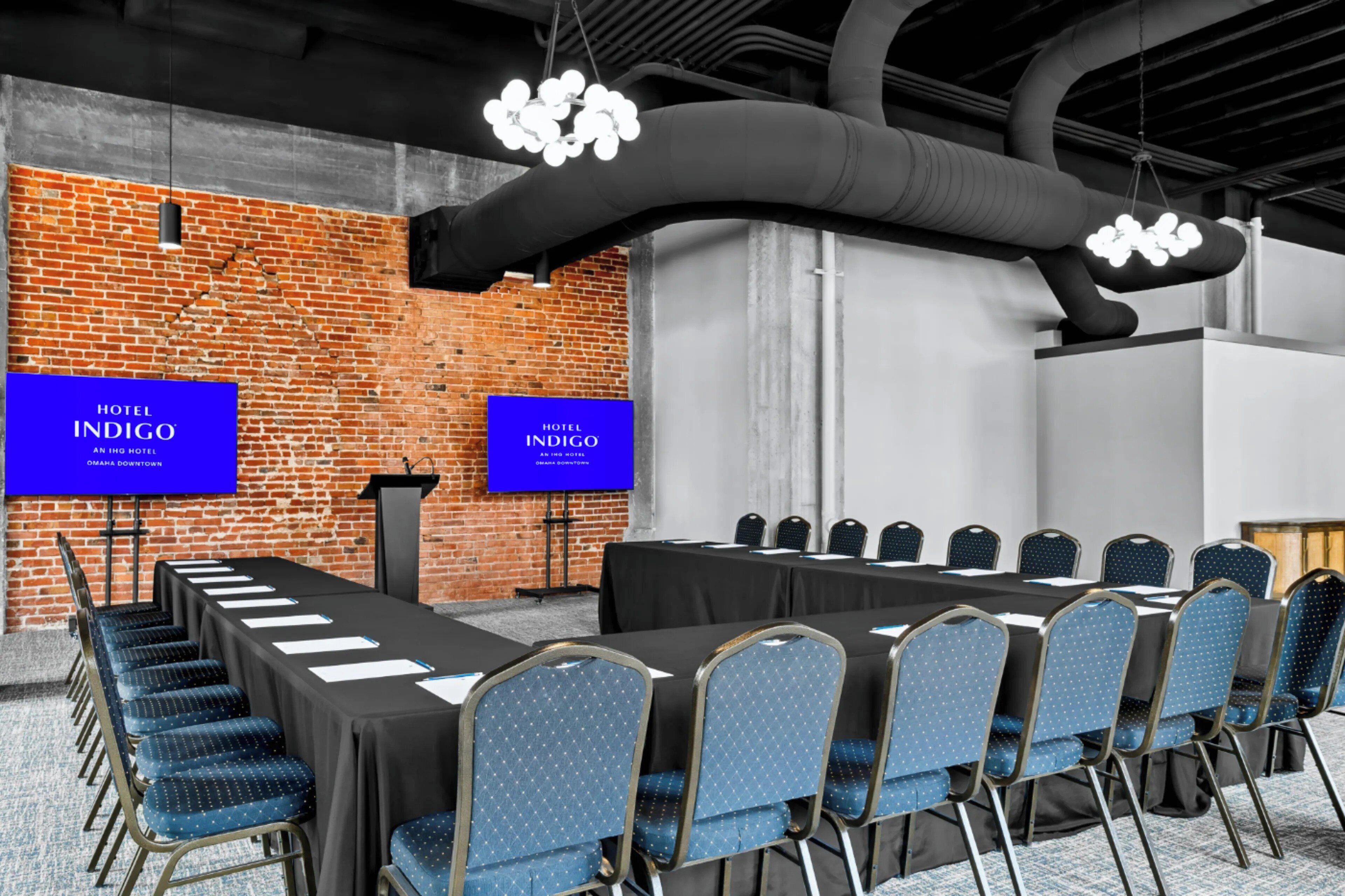 A conference room with a long table, blue chairs, and two screens displaying 'HOTEL INDIGO.'