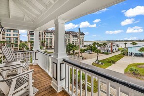 A balcony with wooden flooring and white railings, overlooking a residential area with multiple apartment buildings and a clear sky. - Seaport Village 1473 - 2nd row community in Garden City Beach! (Garden City Beach)