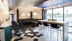 A modern restaurant with a long counter, black and white hexagonal floor tiles, and large windows offering a view of the street. - Hotel Denim (Greensboro)