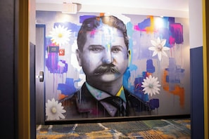 A hallway with a mural of a man, a sign indicating 'Stairs & Exit,' and a patterned carpet. - Hotel Denim (Greensboro)
