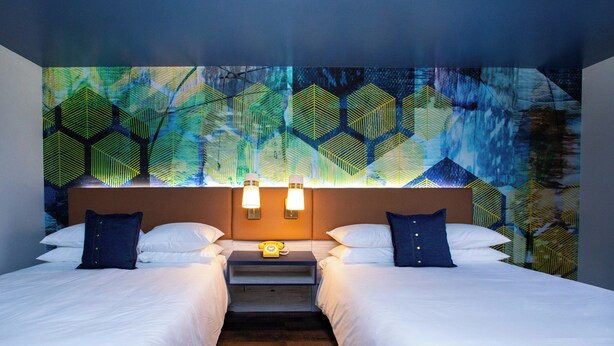 A hotel room with two beds, a nightstand, and a wall with a geometric pattern. - Hotel Denim (Greensboro)