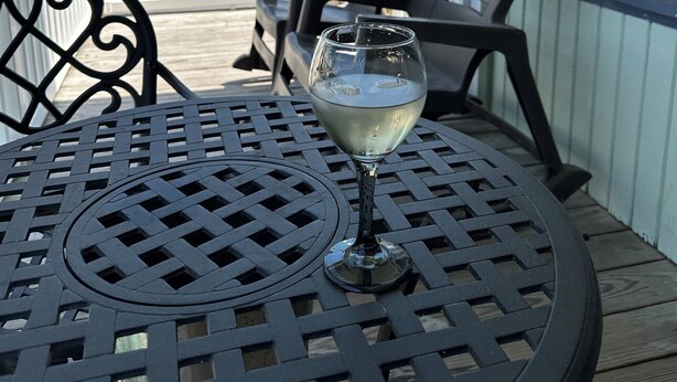 Una copa de vino blanco sobre una mesa de metal con diseño de celosía, dos sillas negras, una terraza de madera y un edificio con fachada azul y blanca al fondo.