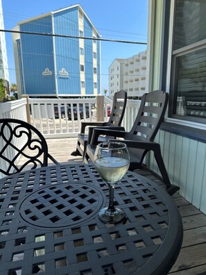 A glass of white wine on a metal table with a lattice design, two black chairs, a wooden deck, and a building with blue and white exterior in the background. - Sunset Square L - 2nd Row in Garden City Beach (Garden City Beach)