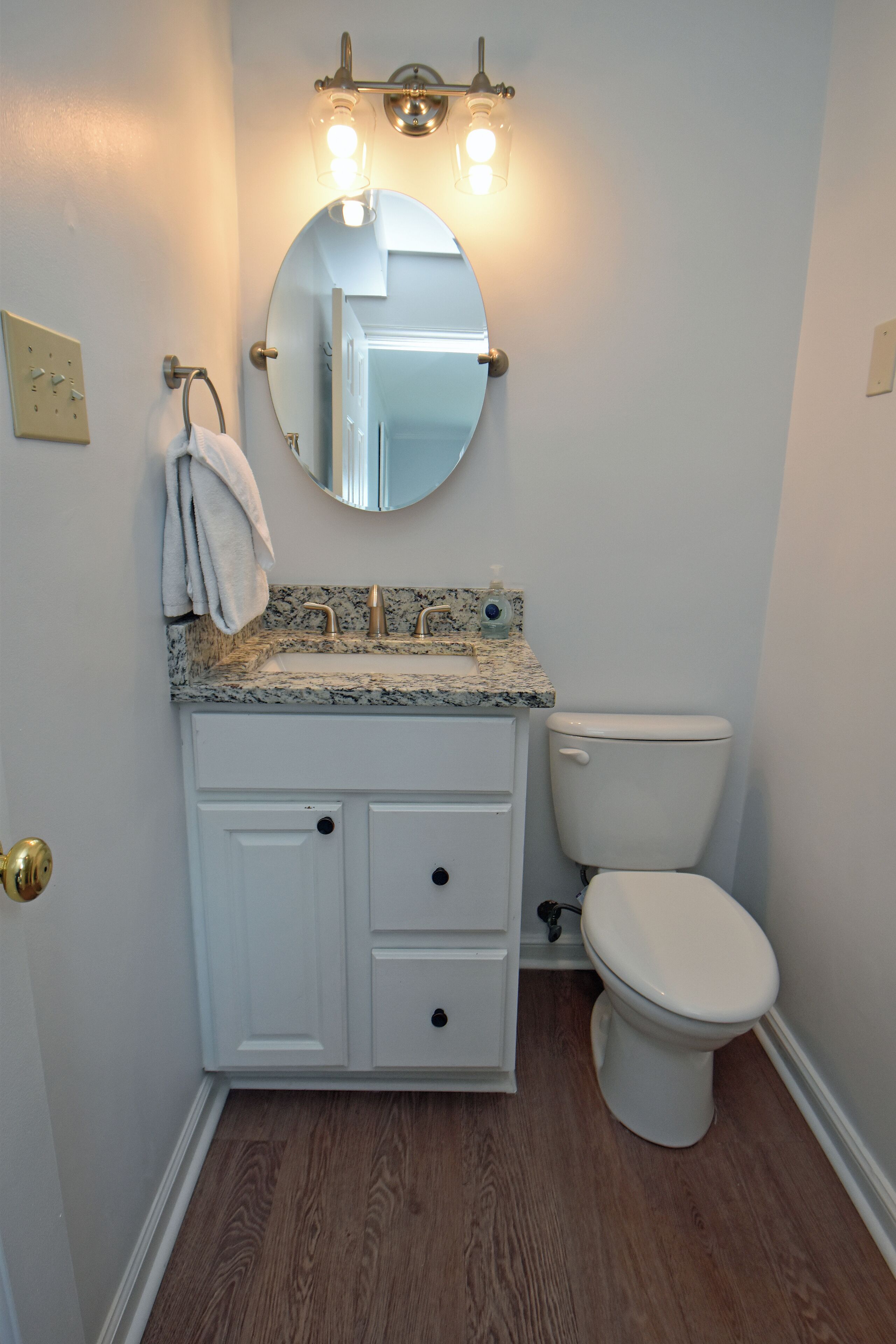 A bathroom with a white vanity, a round mirror, a white toilet, and a wooden floor.