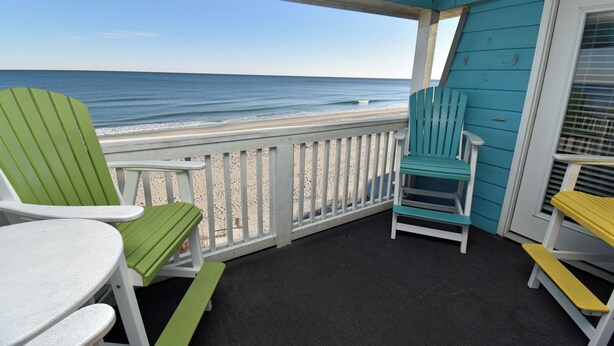 Een balkon met uitzicht op het strand, met groene en witte Adirondack-stoelen en een kleine tafel.
