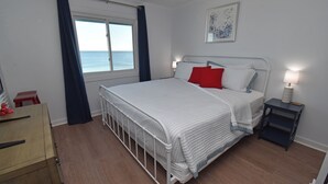 Een slaapkamer met een bed, nachtkastje, dressoir, raam met gordijnen en een schilderij aan de muur.