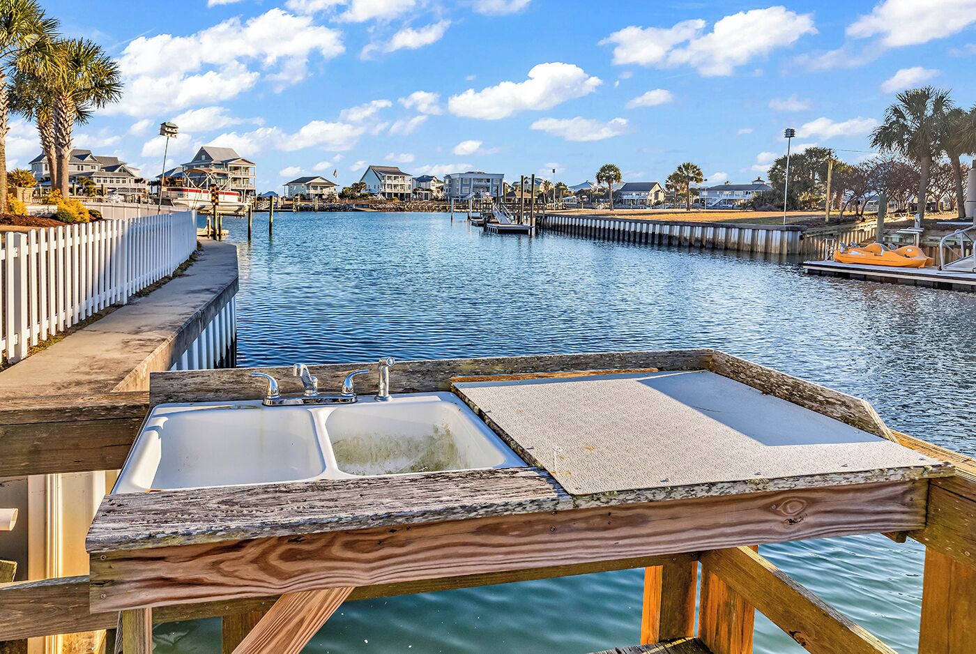 A waterfront view with a wooden dock, a white fence, and a row of houses in the background.