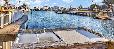 A waterfront view with a wooden dock, a white fence, and a row of houses in the background.