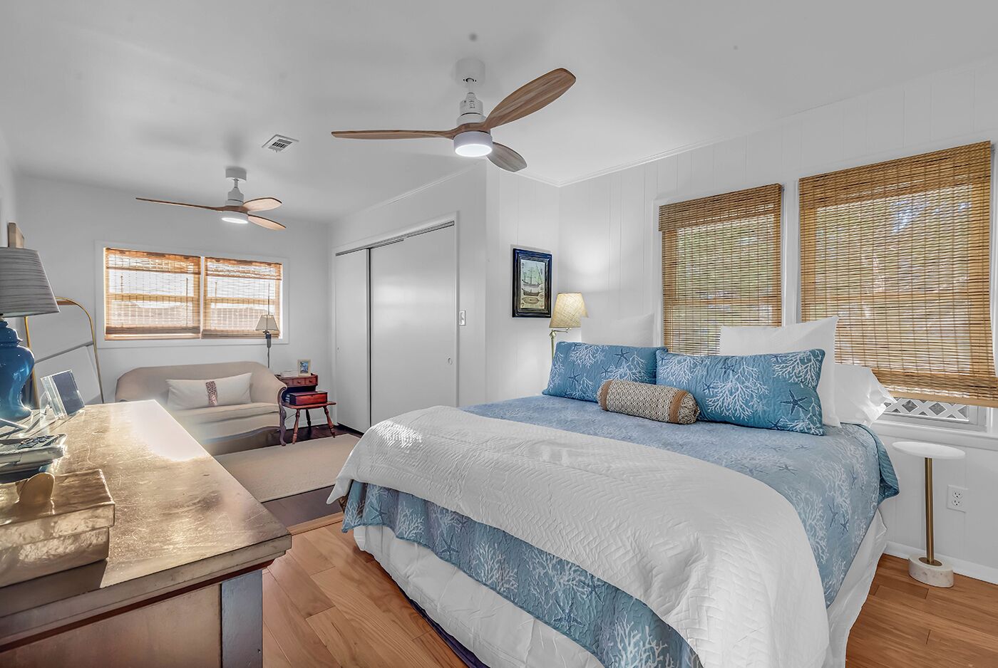A bedroom with a bed, two ceiling fans, a desk with a lamp, a chair, and a window with blinds.