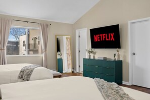 Ein Schlafzimmer mit zwei Betten, einem Fernseher, der 'NETFLIX' zeigt, einem Schminktisch und einem Fenster mit Vorhängen.