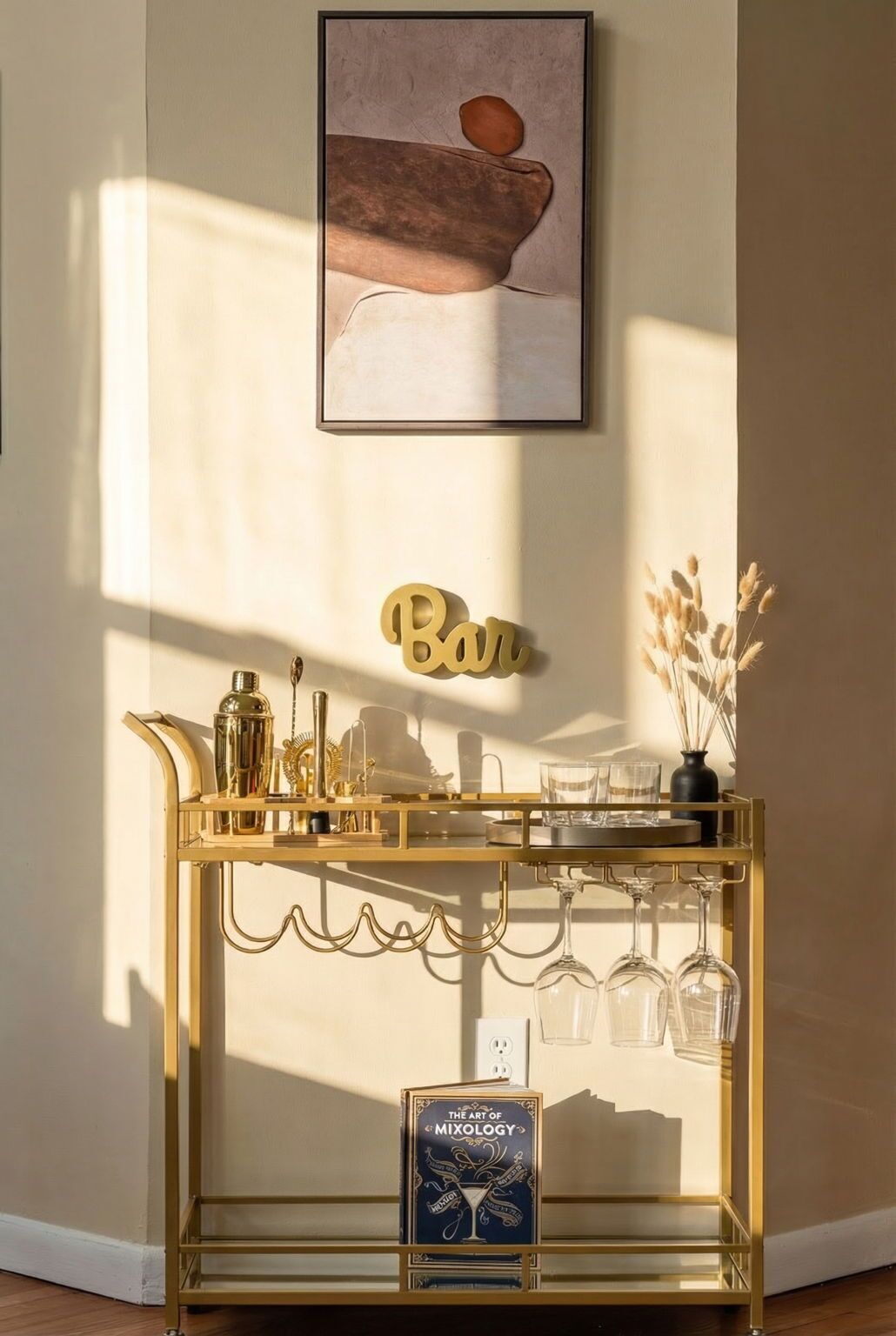 Ein goldfarbener Barwagen mit Glasware, einem Cocktail-Shaker und einem Buch mit dem Titel „The Art of Mixology“ vor einer Wand, über der ein gerahmtes Kunstwerk hängt.