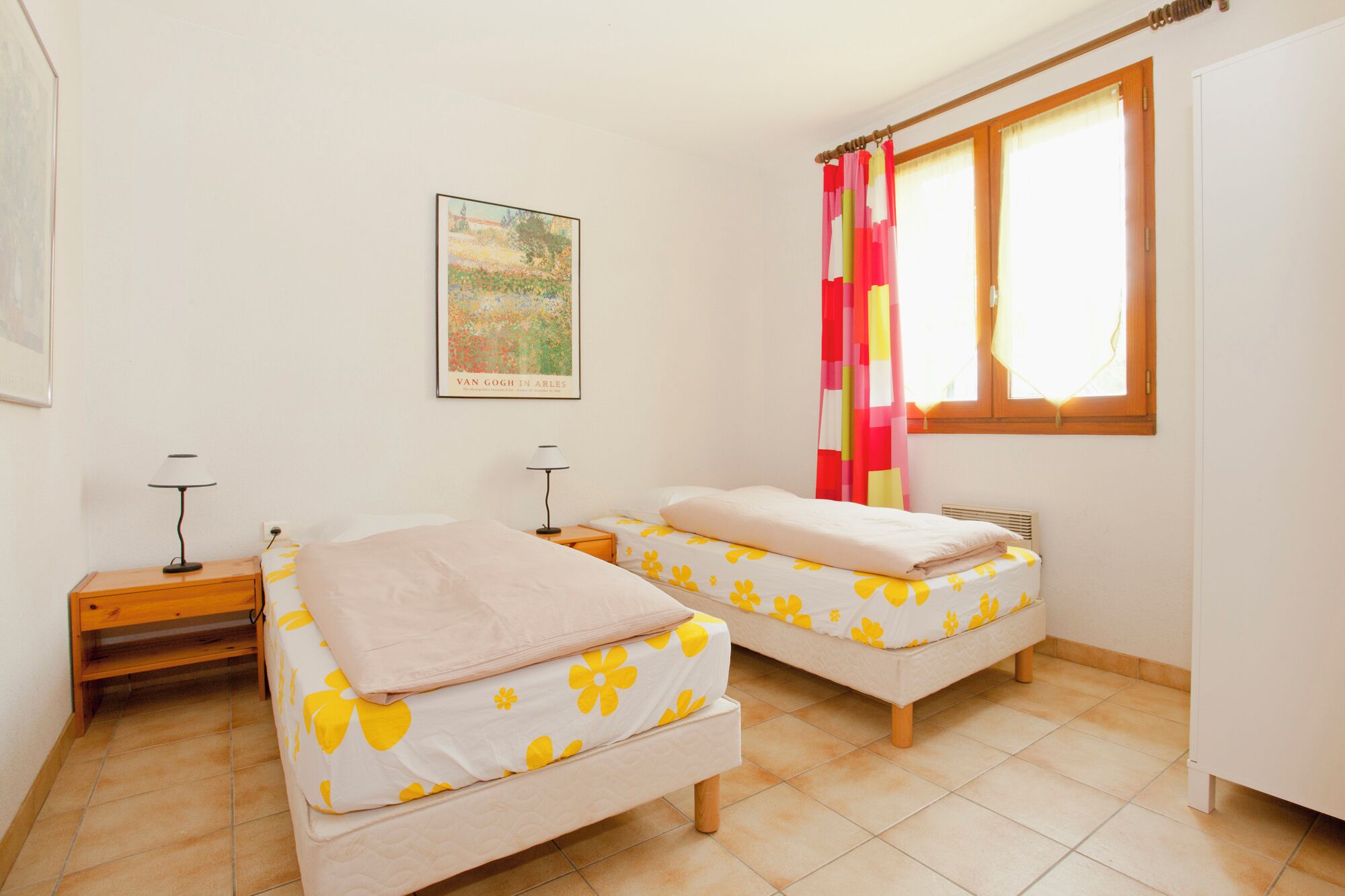 Una habitación con dos camas, una ventana con cortinas, un cuadro colgado en la pared y una mesita con lámpara.