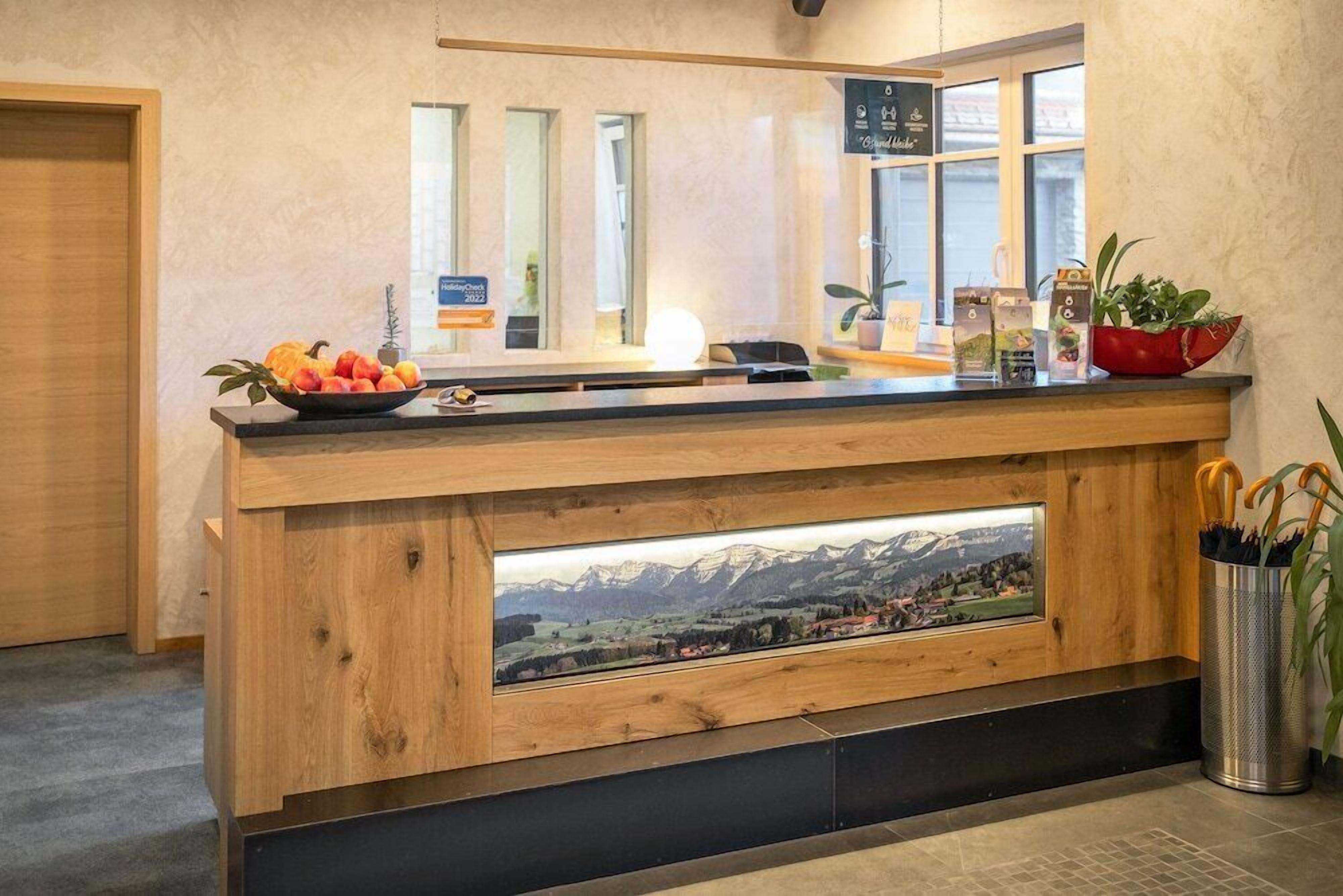 Un comptoir d’accueil avec un revêtement en bois, un panier de fruits, une plante en pot et une vue sur un paysage montagneux par la fenêtre.