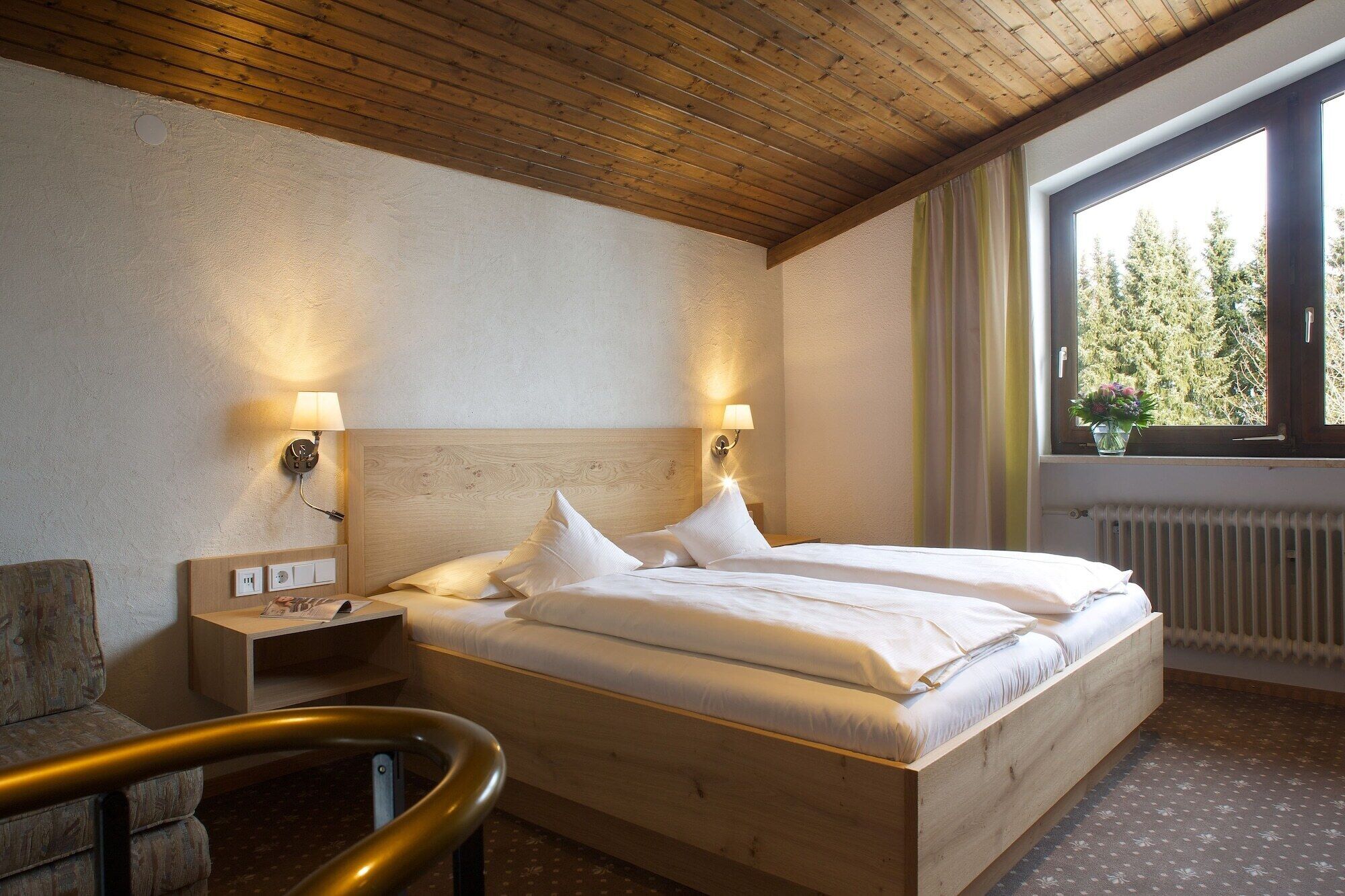 Ein Hotelzimmer mit einem hölzernen Bett, zwei Nachttischlampen, einem Nachttisch, einem Fenster mit Vorhängen und einer Heizung.