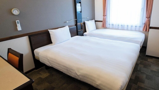 Habitación de hotel con dos camas individuales, un escritorio, una silla, una ventana con cortinas y un termostato en la pared.