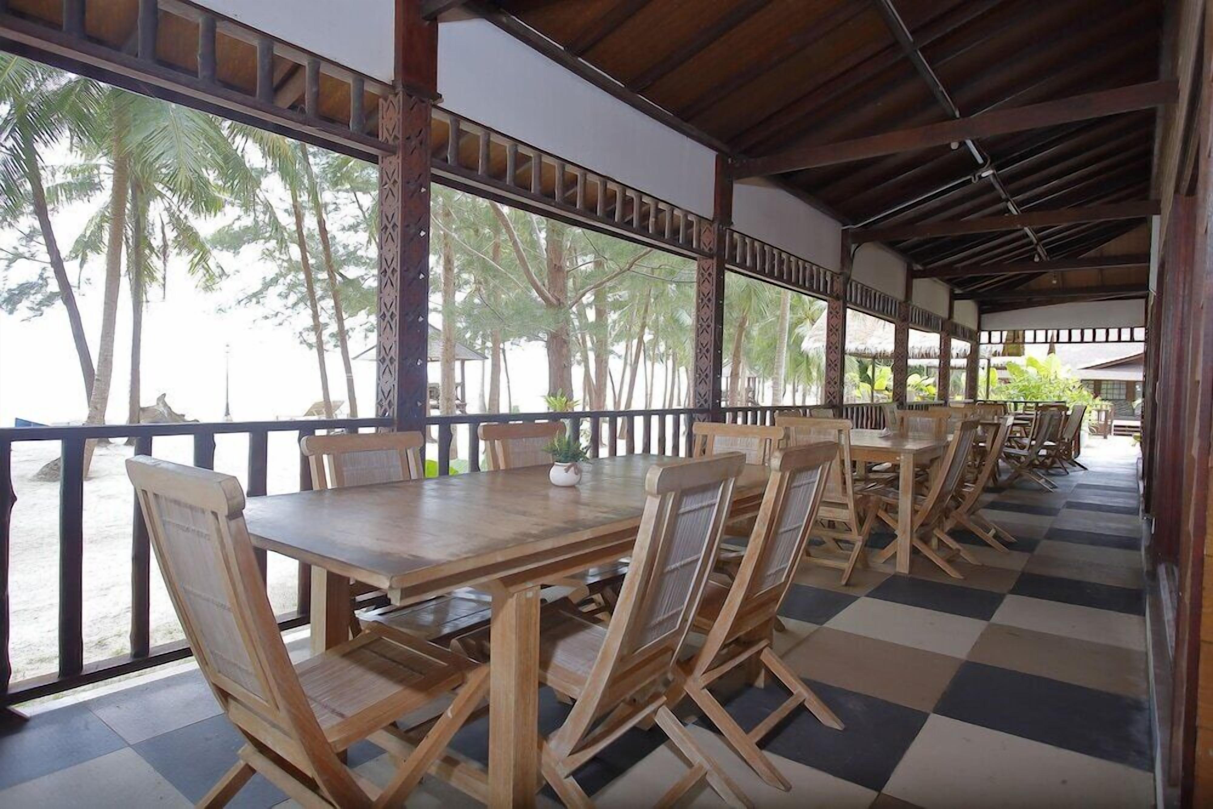 Un espace extérieur abrité, aménagé pour les repas, avec des tables et des chaises en bois, de grandes fenêtres et un sol à carreaux.