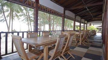 Un espace extérieur abrité, aménagé pour les repas, avec des tables et des chaises en bois, de grandes fenêtres et un sol à carreaux.
