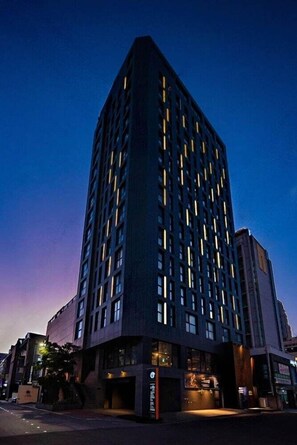 A tall, modern building with a corner entrance and illuminated windows. - Hotel Alice and Trunk (Jeju City)