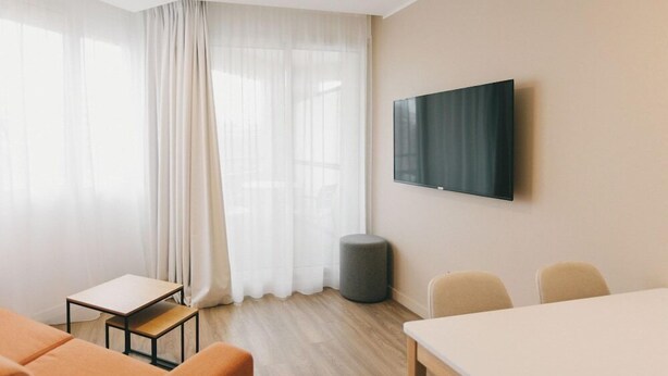 Una habitación moderna y bien iluminada, con un gran ventanal, un televisor de pantalla plana montado en la pared, un pequeño taburete redondo y una mesa con sillas.