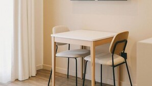 Una habitación con una televisión de pantalla plana en la pared, una mesa de madera con dos sillas y un gran ventanal con cortinas blancas.