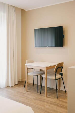 A room with a flat-screen TV mounted on the wall, a wooden table with two chairs, and a large window with white curtains. - Appart Hotel Marianne (Montpellier)