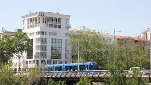 Un edificio moderno con el nombre 'MIRANOVE' en la fachada, un tren azul pasando y un puente con vegetación en primer plano.