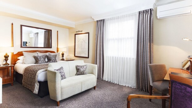 A hotel room with a bed, a sofa, a chair, a desk, a lamp, a mirror, and a window with curtains. - Brandshatch Place Hotel and Spa (Longfield)