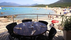 Un restaurant situé en bord de mer, avec une table dressée pour deux, offrant une vue sur une plage animée, remplie de monde et de bateaux.
