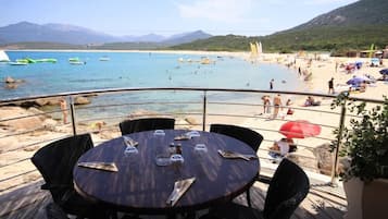 Un restaurant situé en bord de mer, avec une table dressée pour deux, offrant une vue sur une plage animée, remplie de monde et de bateaux.