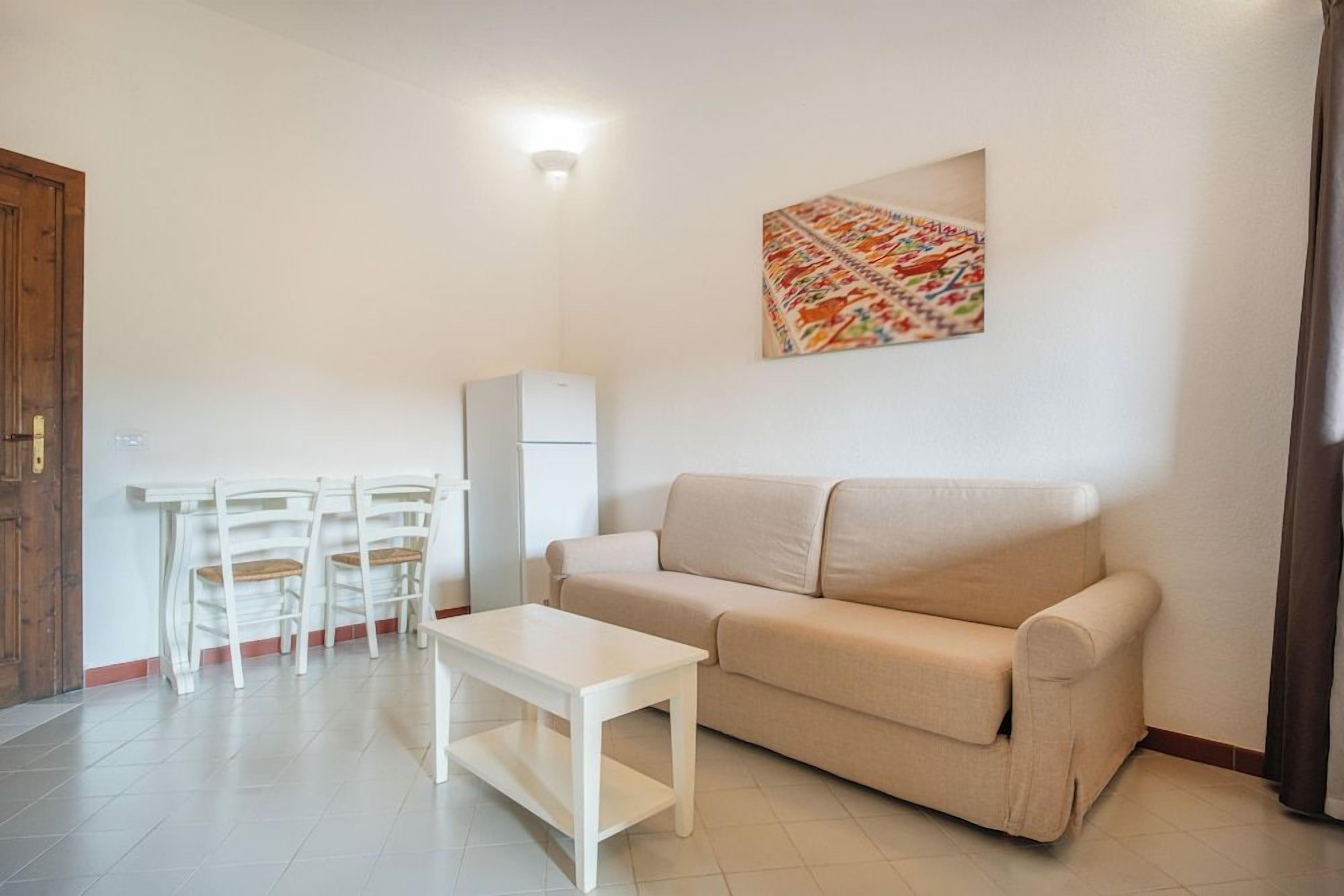 A living room with a beige sofa, a white coffee table, and a small dining area with white chairs and a refrigerator.