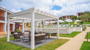 A covered outdoor seating area with wicker furniture and umbrellas, a pathway, and a building with balconies in the background. - Residence Costa Serena (Palau)