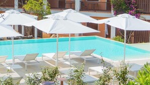 A pool area with white umbrellas and lounge chairs, surrounded by greenery and a building with balconies. - Residence Costa Serena (Palau)